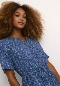 Modra cvetna obleka s kratkimi rokavi, zapenjanjem na gumbe in vezanjem v pasu. Ima vzorec majhnih belih in svetlo modrih cvetov na blagu.