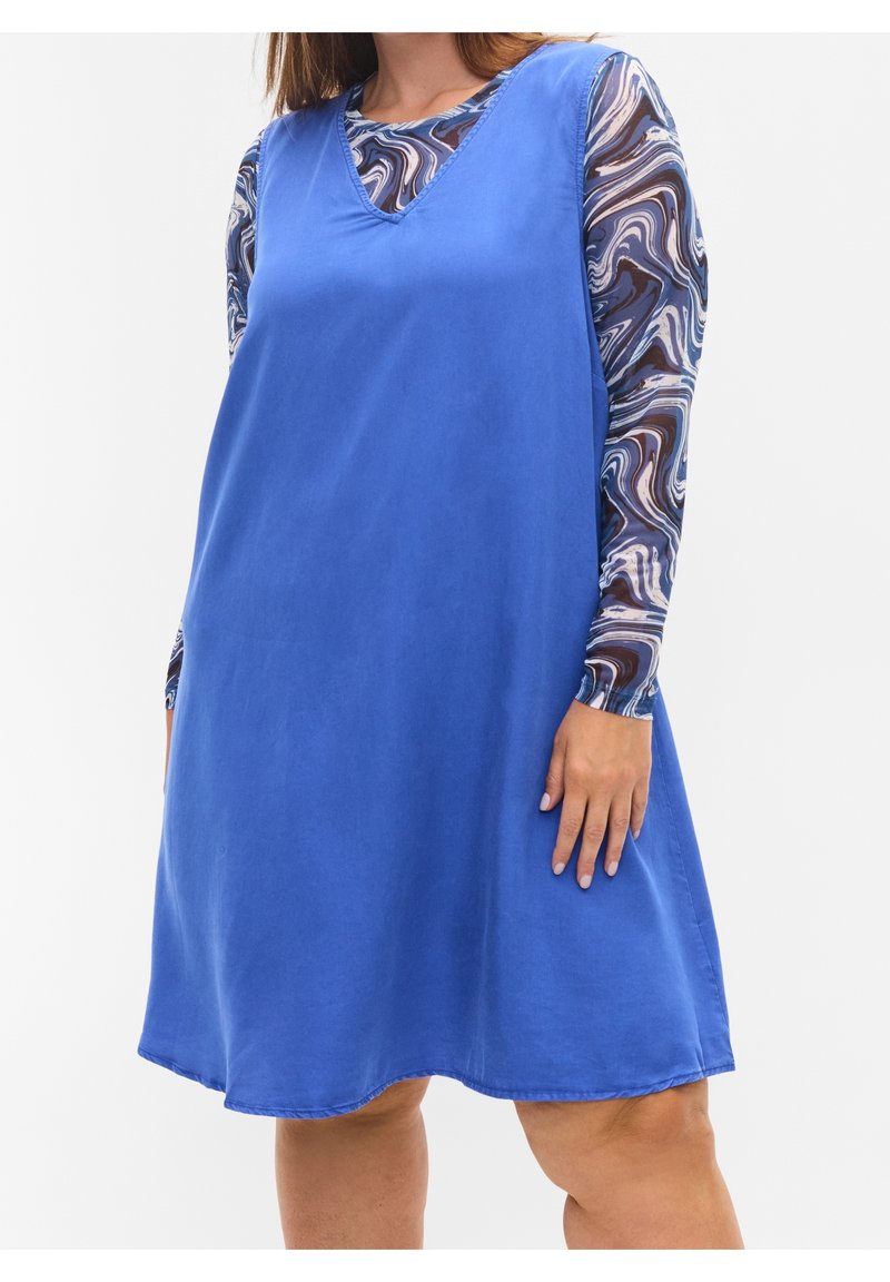 Blå ärmlös klänning med v-ringning och en lös passform, ihop med en mönstrad långärmad topp med virvlar i brunt och kräm.
