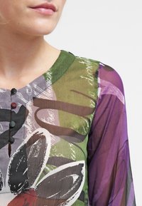 Blusa trasparente caratterizzata da una combinazione di colori verdi e viola, con motivi floreali e una chiusura con bottoni. Collo rotondo e maniche lunghe.