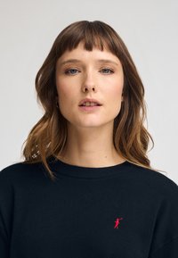 Suéter de cuello redondo en color marino con puños acanalados, un corte relajado y un pequeño logo rojo en la parte izquierda del pecho. Textura suave en toda la prenda.