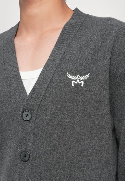 Grå cardigan gjord av mjukt tyg, med V-hals, tre svarta knappar och ett vitt broderat emblem på vänster bröst.