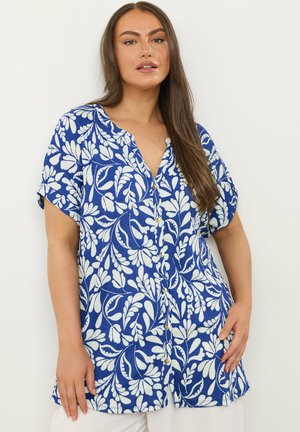 Frau mit langen braunen Haaren, die ein blaues Kurzarmhemd mit weißen Blumenmustern trägt, vor einem einfarbigen Hintergrund stehend.