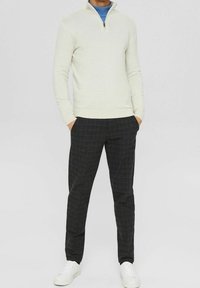 Helles beiges Zip-up Sweatshirt, langer Ärmel, gerippte Bündchen. Dunkelkarierte Hose, figurbetonter Schnitt mit Taschen. Weiße Sneaker runden den Look ab.