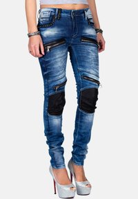 Jeans skinny in denim blu con molte tasche con zip, toppe nere sulle ginocchia e borchie decorative sul cinturino, completati da un lavaggio sbiadito.
