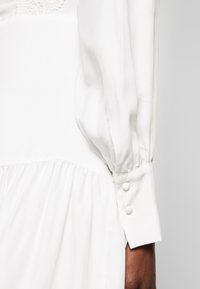 Biała sukienka z teksturowanym koronkowym górnym fragmentem i bufkami. Posiada marszczoną talię i mankiety z dwoma guzikami.