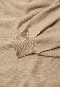 Tissu en maille beige avec détail visible de bord-côte côtelé, révélant une texture douce et de délicats plis sur la manche d'un pull.