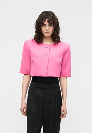 Vrouw met krullend haar staat in een felroze cropped jasje met twee knopen aan de voorkant en een hoge taille zwarte broek.