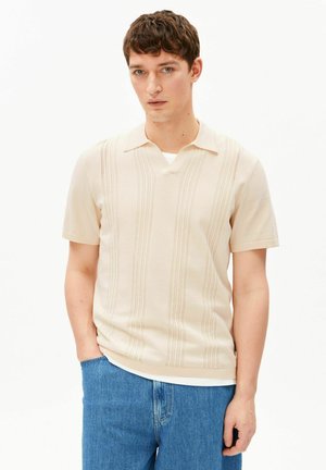 Jonge man draagt een beige korte mouwen gebreide polo met verticale patronen en een blauwe spijkerbroek, staand met één hand in de zak.