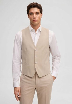 Giovane uomo con capelli ricci che indossa un gilet beige e pantaloni abbinati sopra una camicia bianca, in piedi con una mano in tasca.
