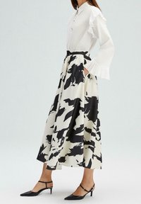 Femme portant une longue jupe noire et blanche à motif abstrait, une blouse blanche à manches volantées et des chaussures noires à talons bas.