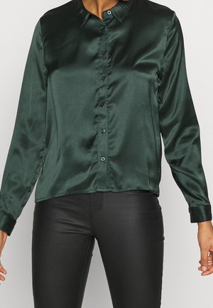 Woman wearing a dark green satin long-sleeve button-up shirt and black fitted pants, standing against a plain background.