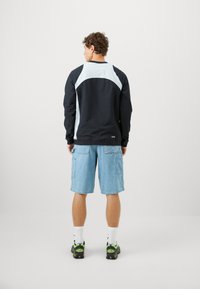 Langærmet skjorte i sort og lyseblå med et buet design, parret med lyseblå cargo shorts og sorte sneakers med grønne accenter.