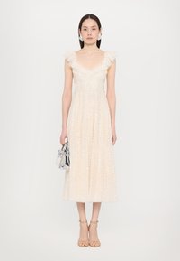 Beige midikleid mit gerafften Schultern, verziert mit Pailletten, mit einem taillierten Oberteil und fließendem Rock. Begleitet von einer silbernen Tasche.
