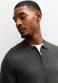 Dunkelgrünes Poloshirt aus strukturiertem Strick, mit einem Reißverschlusskragen und kurzen Ärmeln, getragen über einem weißen Unterhemd.