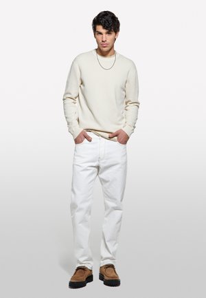 Pull en texture couleur crème, jean blanc et chaussures brunes avec semelles noires. Tenue décontractée, mannequin se tient les mains dans les poches contre un fond uni.