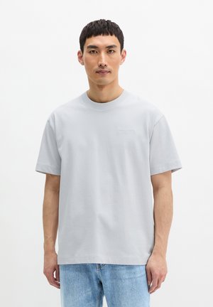 Jeune homme aux cheveux noirs courts portant un t-shirt gris clair à col rond et un jean bleu clair, debout devant un fond blanc uni.