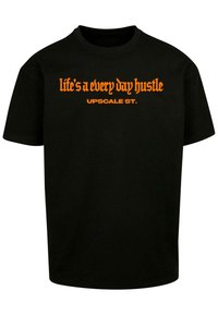 Black cotton t-shirt featuring orange text that reads “life's a every day hustle” and “UPSCALE ST.” in distinct fonts. Short sleeves.