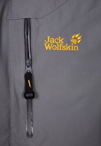 Šedá bunda s kapsou na zip se černým jezdcem, žlutým vyšitým logem "Jack Wolfskin" a detailním potiskem tlapky na látce.