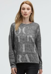 Grauer Pullover mit langen Ärmeln, weichem Material, versehen mit großen hellgrauen Schriftzügen. Kombiniert mit schwarzen Jeans mit Nietenakzenten.