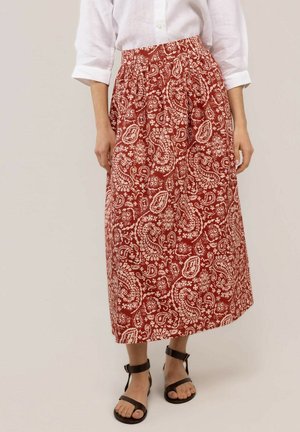 Mujer con blusa blanca y falda midi roja con estampado paisley crema, combinada con sandalias planas negras con tira en el tobillo.