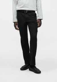 Mustad püksid kohandatud lõikega, valmistatud tekstuurse kangaga, esitaskutega, kantud halli pikkade varrukatega särke ja mustade kingadega.