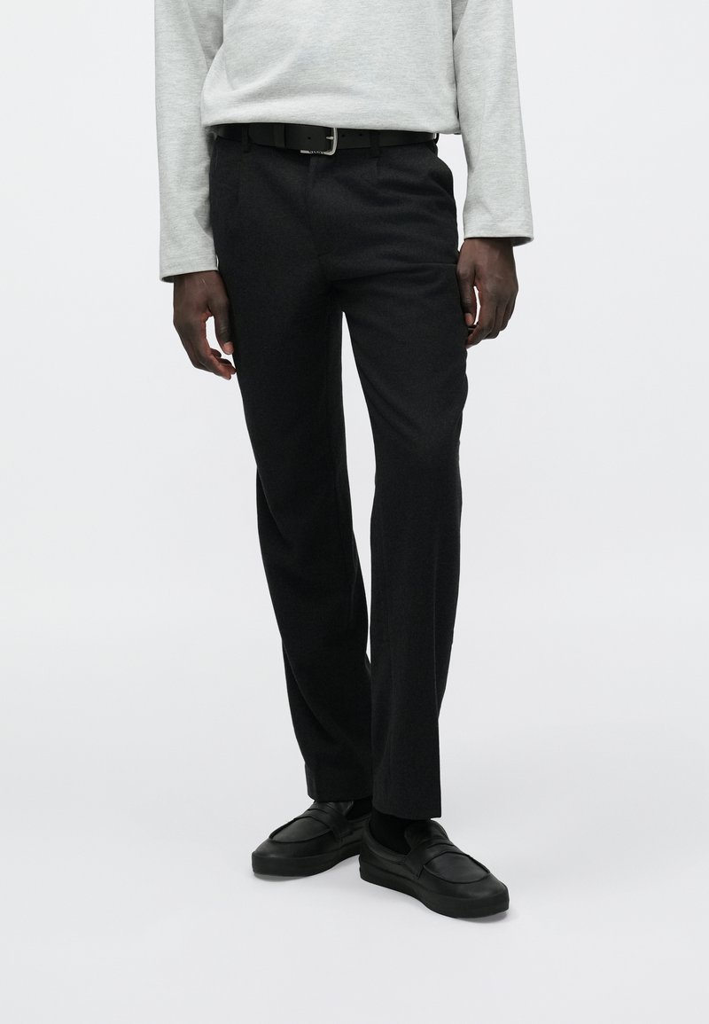 Mustad püksid kohandatud lõikega, valmistatud tekstuurse kangaga, esitaskutega, kantud halli pikkade varrukatega särke ja mustade kingadega.