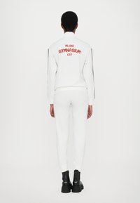 Witte trainingsoutfit met een aansluitende pasvorm, voorzien van een rode borduring "MILANO GYMNASIUM EA7" op de achterkant, gecombineerd met zwarte chunky laarzen.