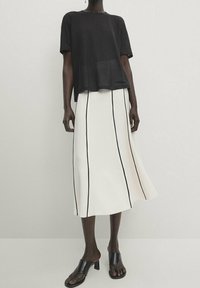 Schwarzes kurzärmeliges Oberteil kombiniert mit einem weißen Midirock mit vertikalen schwarzen Streifen, ergänzt durch schwarze offene Mules.