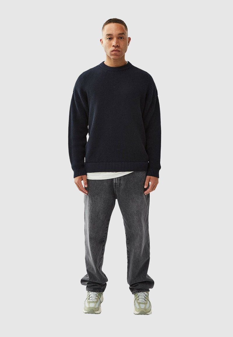 Dark navy knitted sweater with a crew neck, paired with loose grey jeans and green-tinted sneakers. Textured fabric, relaxed fit.
