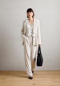 Light striped suit consisting of a tailored blazer and matching trousers, paired with a buttoned vest. A black handbag completes the look.
