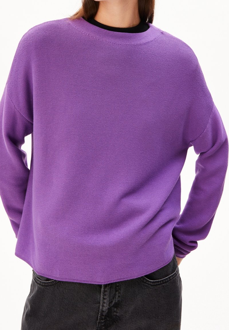Personne portant un pull violet ample à manches longues sur un haut noir, associé à un pantalon gris foncé, montrée du cou aux hanches.