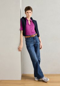 Kortermet magenta polo med knapper, kombinert med høy midje blå rette jeans og lysegrå sko. Marineblå genser drapert over skuldrene.
