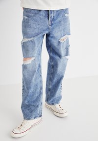 Jean en denim bleu clair avec des déchirures multiples, coupe décontractée et design à jambe droite. Portés avec des baskets en toile blanches.