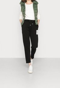 Schwarze Jogginghose mit weißem Logo-Patch, Kordelzugbund und zulaufenden Hosenbeinen. Kombiniert mit einer grünen Steppweste und weißen Turnschuhen.