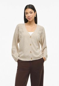 Žena s rovnými černými vlasy, která má na sobě zlaté kruhové náušnice, béžový kardigan s knoflíky s květinovými vzory, bílý top a tmavě hnědé kalhoty.