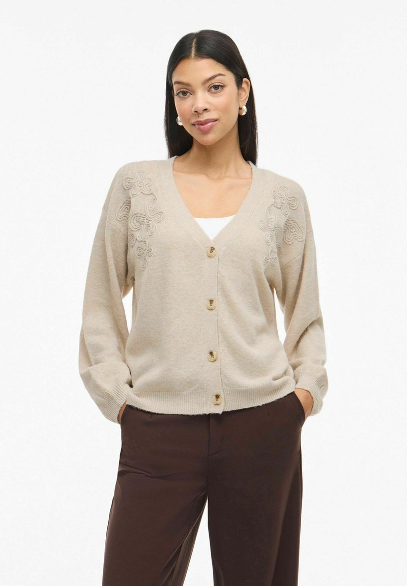 Žena s rovnými černými vlasy, která má na sobě zlaté kruhové náušnice, béžový kardigan s knoflíky s květinovými vzory, bílý top a tmavě hnědé kalhoty.