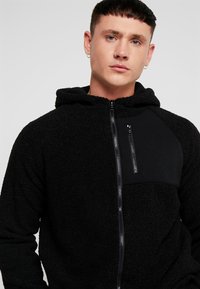 Sort lynlås hoodie lavet af tekstureret fleece med en glat sektion på brystet. Har en frontlynlås lomme og justerbar hætte.