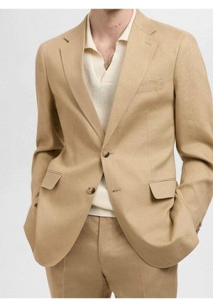 Mand iført en beige jakkesæt med en kravecremestriktrøje under, hænderne stoppet i jakkelommerne, beskåret ved nakke og lår.