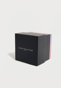 Μαύρο ορθογώνιο κουτί με το λογότυπο "Tommy Hilfiger Watches" σε λευκό και μια μικρή σημαία κόκκινου, λευκού και μπλε σε απλό λευκό φόντο.