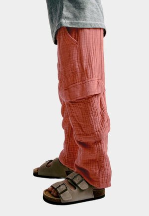 Losse, koraalkleurige broek van gestructureerde stof, met zijzakken en een omgeslagen zoom, gecombineerd met grijze sandalen met dubbele gespen.