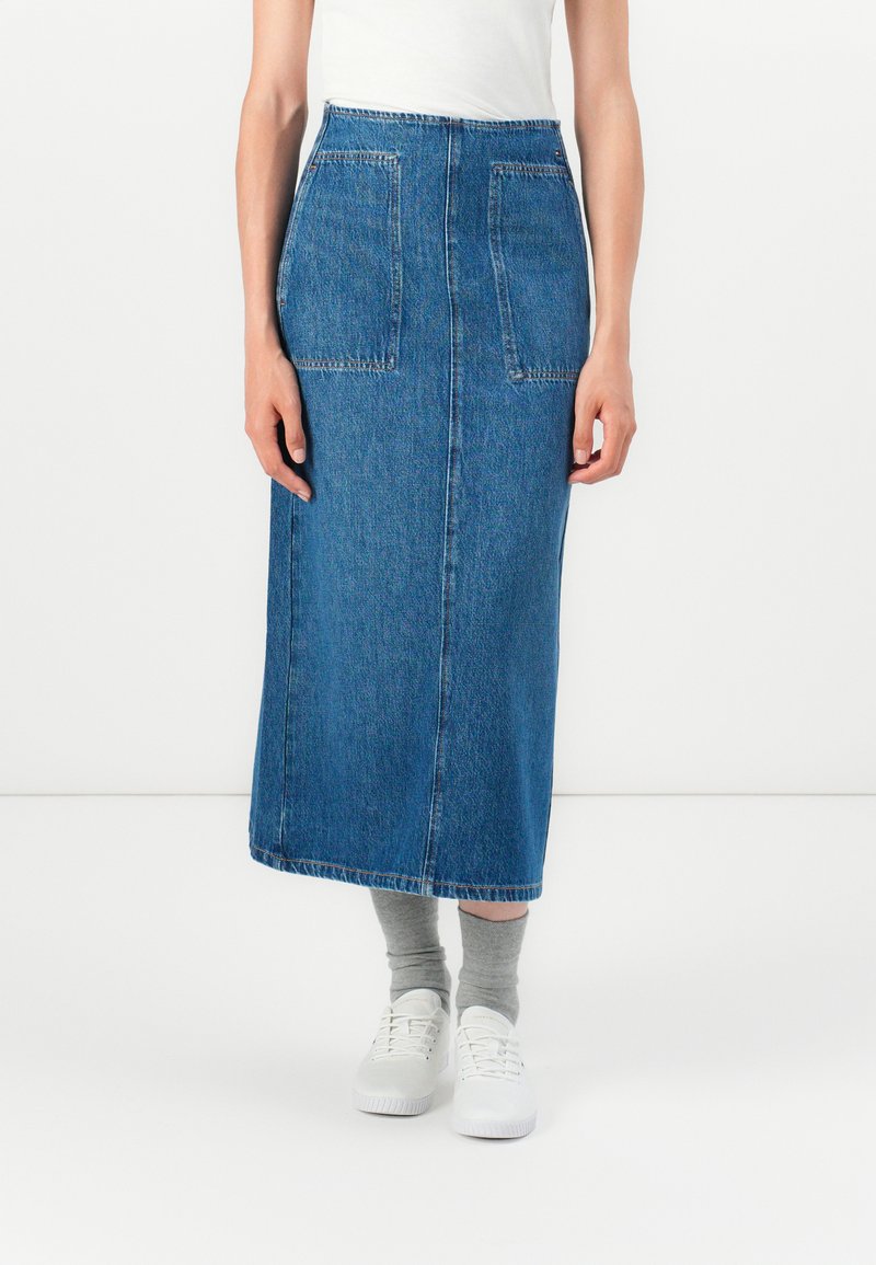 Denim midi sukňa s dvoma prednými vreckami, surovým lemom a priliehavým pásom. Nosená s bielymi teniskami a sivými ponožkami.
