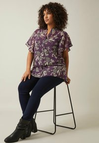 Lila Bluse mit floralen Motiven, kurzen, fließenden Ärmeln und einer durchgehenden Knopfleiste, kombiniert mit dunkelblauen engen Jeans und schwarzen, hochhackigen Stiefeletten.
