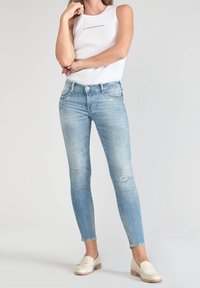 Vaqueros ajustados azul claro con detalles desgastados, acompañados de un top blanco de canalé y mocasines crema, sobre un fondo liso.