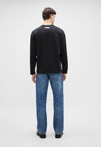 Svart langermet skjorte med rund hals og etikett på baksiden, kombinert med lyseblå rette jeans og svarte sko.