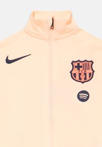 Šviesiai persikų spalvos sportinė striukė su aukšta apykakle, su tamsiai mėlynu Nike logotipo, išsiuvinėtu Barselonos emblemą ir apvaliu Spotify logotipu.