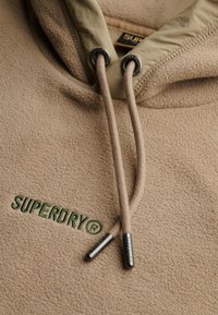 Tan fleece hoodie met een beige capuchon en trekkoorden, met groene "SUPERDRY" borduursels en zilveren hardware accenten.
