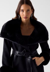Zwarte jas met een brede sjaalkraag van namaakbont, leren accenten en riem. Heeft lange mouwen met bontmanchetten en een knoopsluiting.