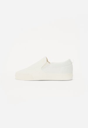 Baskets blanches à enfiler avec un panneau avant texturé, des côtés lisses, et un design minimaliste avec une boucle arrière discrète pour un enfilage facile.