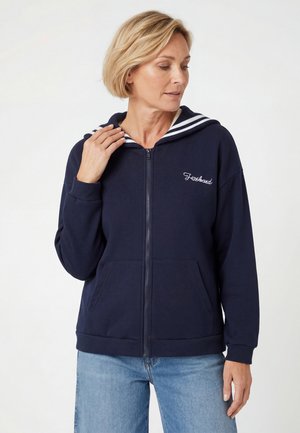Kobieta w granatowej bluzie z kapturem na zamek błyskawiczny z białym paskowanym kołnierzem i jasnoniebieskich dżinsach, patrząca w dół i dotykająca kołnierza jedną ręką.