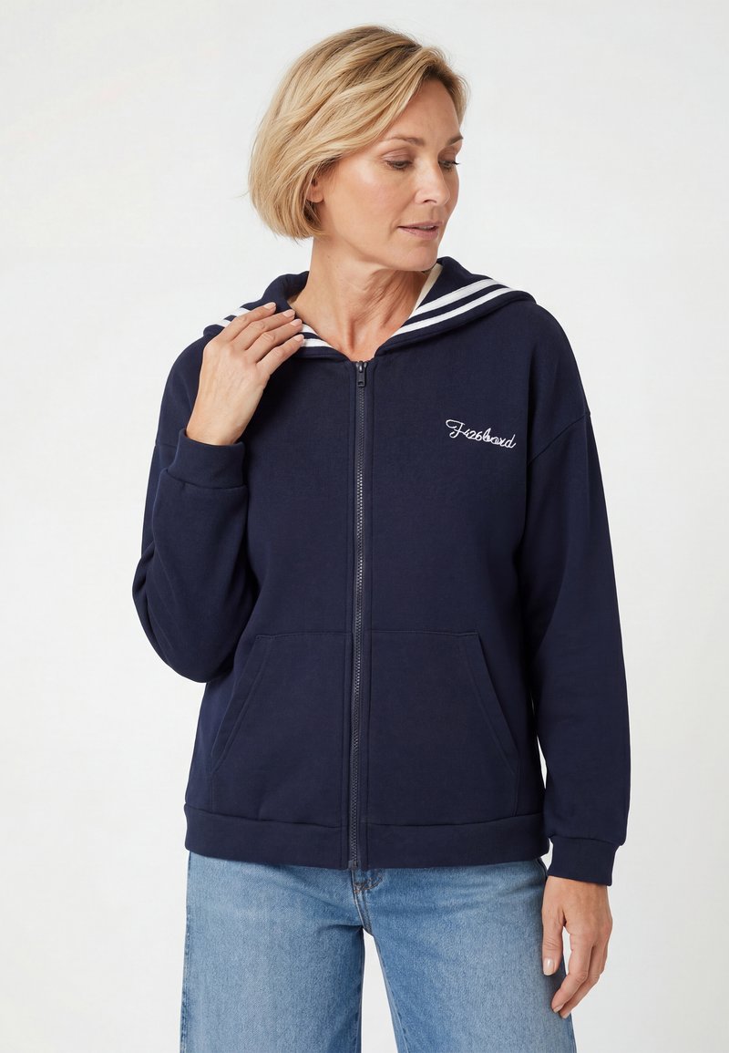 Femme portant un sweat à capuche bleu marine zippé avec un col rayé blanc et un jean bleu clair, regardant vers le bas et touchant le col d'une main.